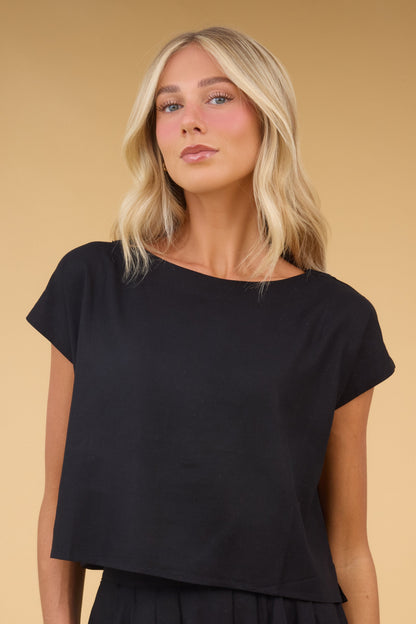Young woman wearing a loose fiting black top with a boat neckline and cap sleeves.