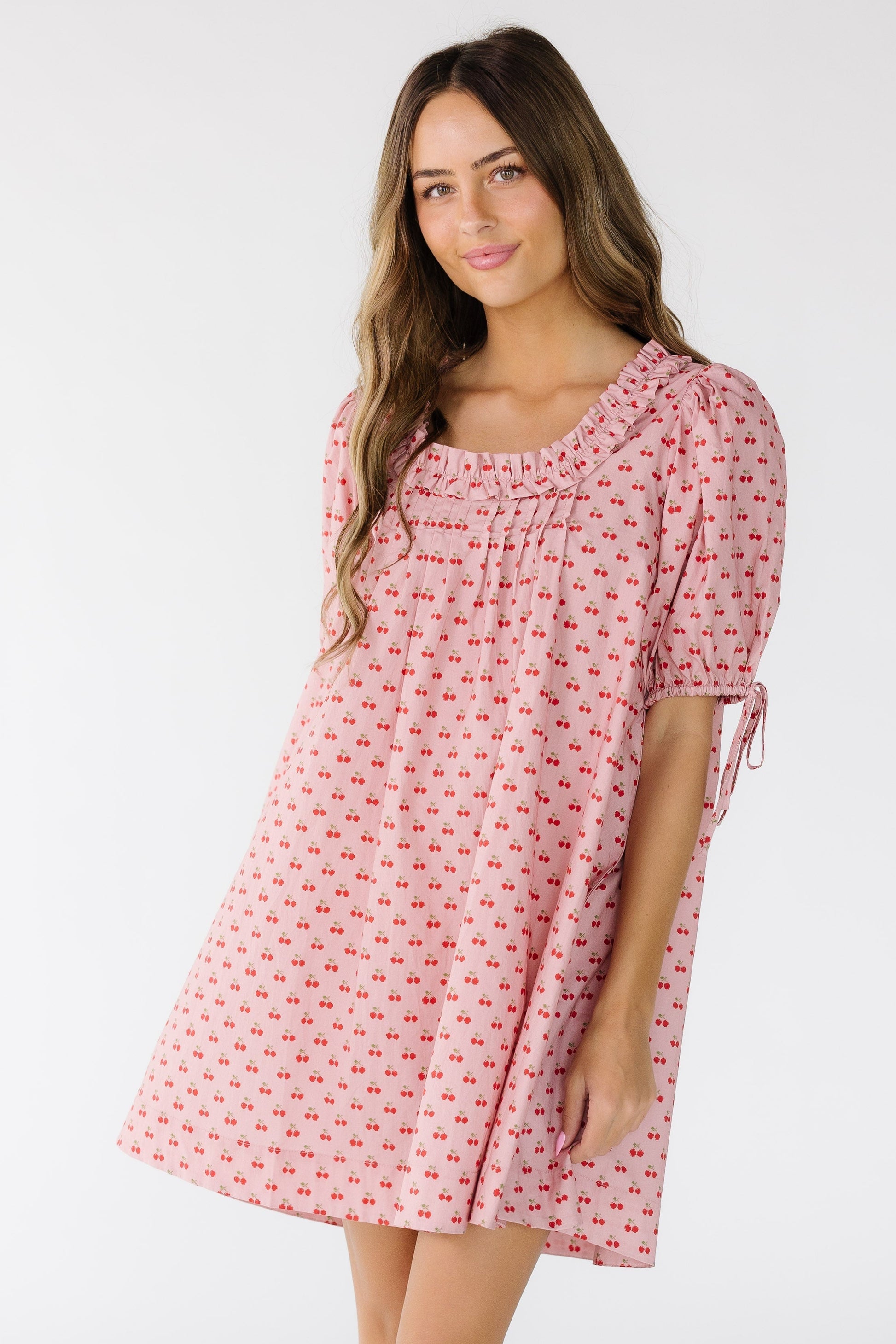 Woman wearing a modest pink cherry print short dress with a square neckline with ruffles and pleats.