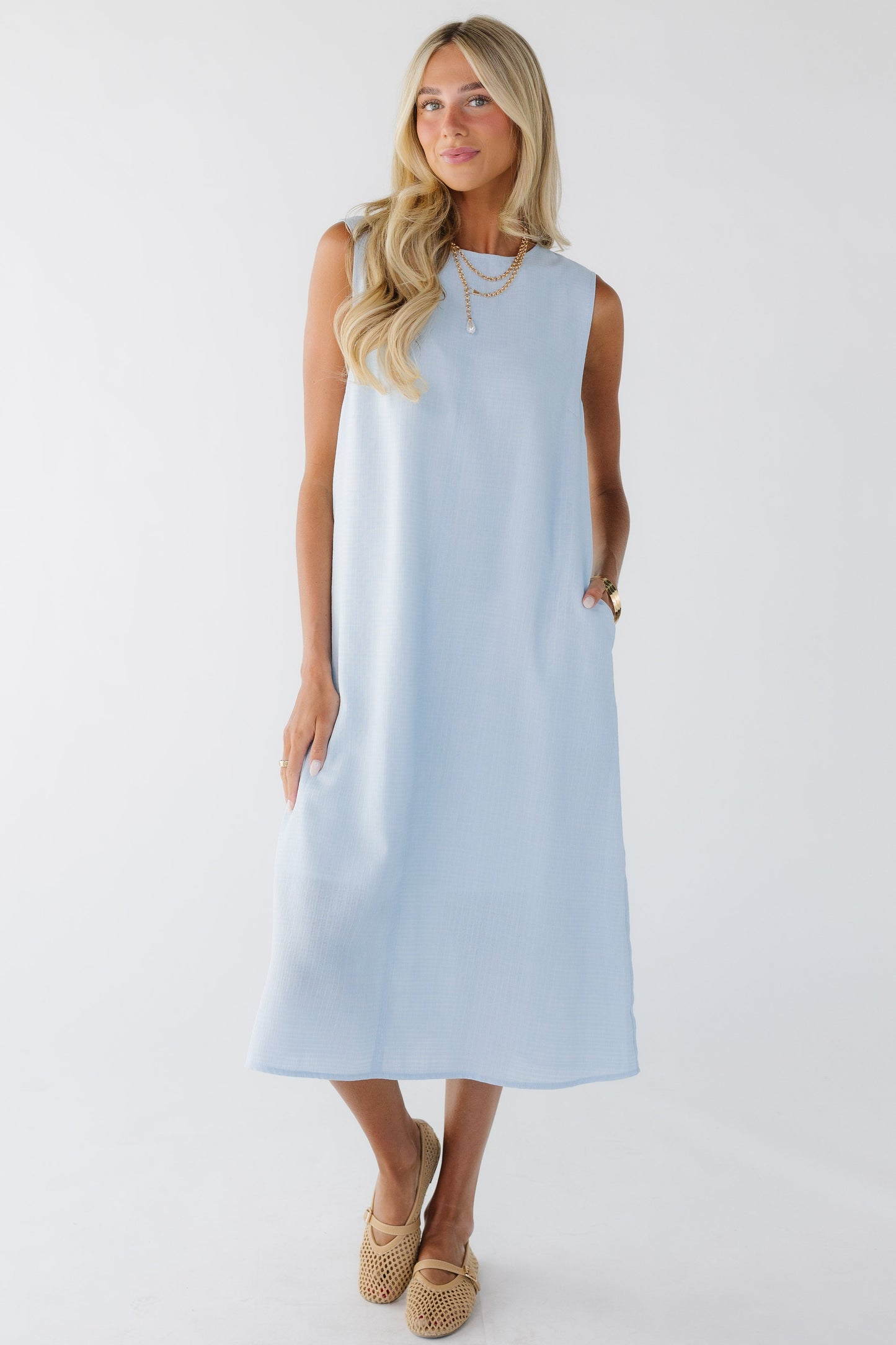 Young woman wearing a sleeveless midi length shift dress with side pockets in light blue.