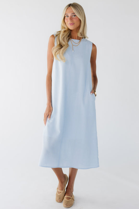 Young woman wearing a sleeveless midi length shift dress with side pockets in light blue.