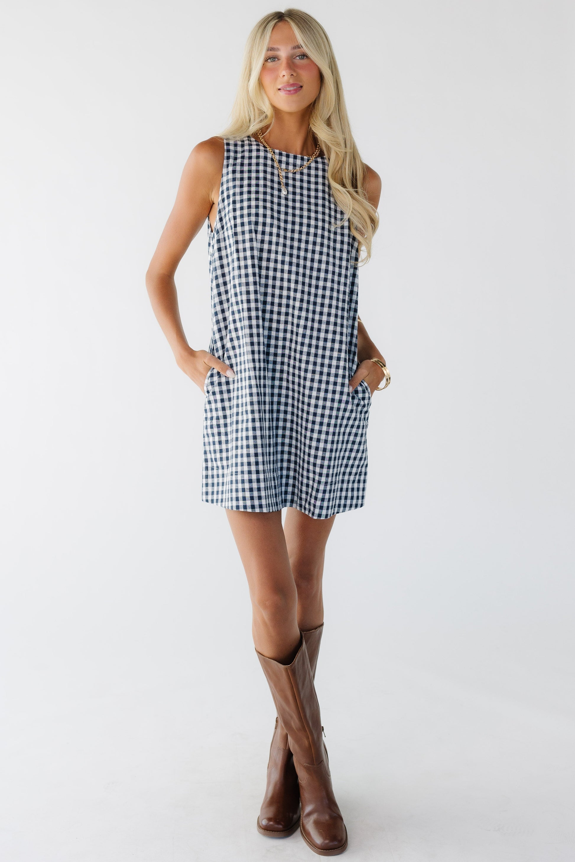 Young woman wearing a navy & white gingham sleeveless shift dress with side pockets.