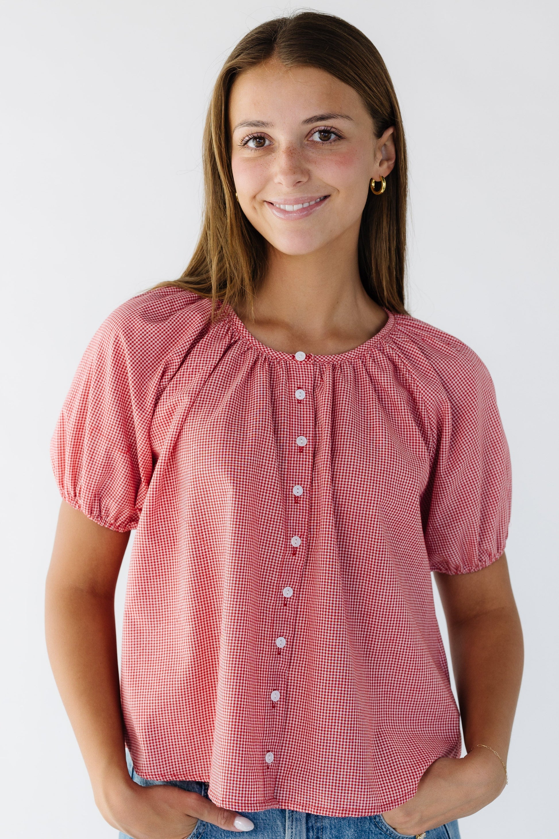 Young woman wearing  a modest loose fitting button down blouse in red gingham with around neckline and short puff sleeves.