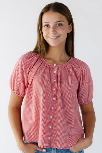 Young woman wearing  a modest loose fitting button down blouse in red gingham with around neckline and short puff sleeves.