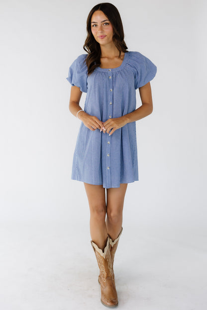 Young woman wearing a navy & white gingham, button down short dress with a scoop neckline and balloon sleeves.