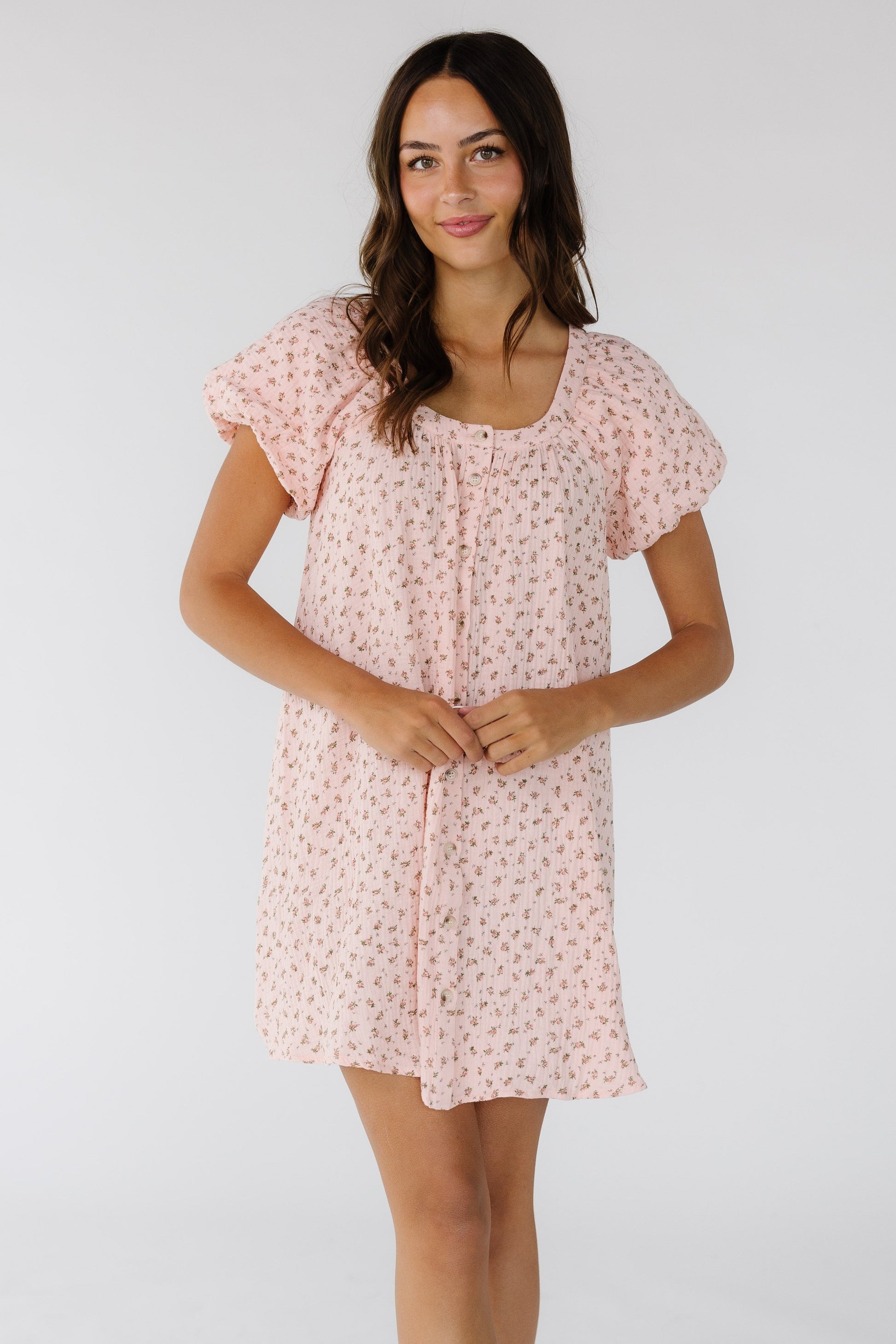 Young woman wearing a button down pink floral short dress with a scoop neckline and short balloon sleeves.