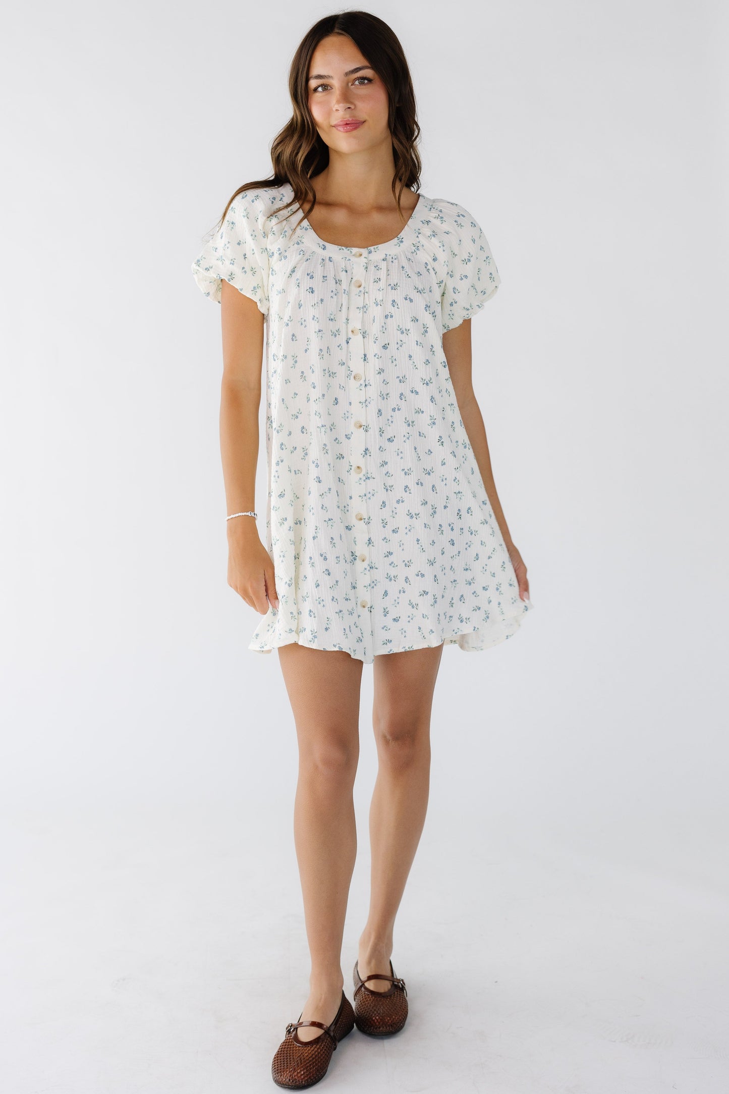 Young woman wearing a button down short dress in white with blue floral print with a scoop neckline.