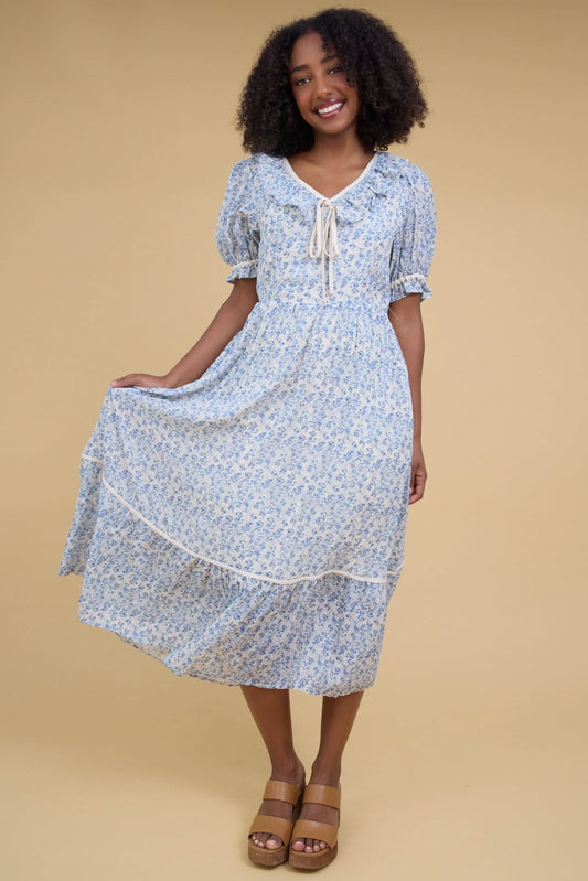 Young woman wearing a modest blue floral midi dress iwth a ruffled V-neckline, short puff sleeves with a ruffle cuff and a full skirt with a wide bottom ruffle.