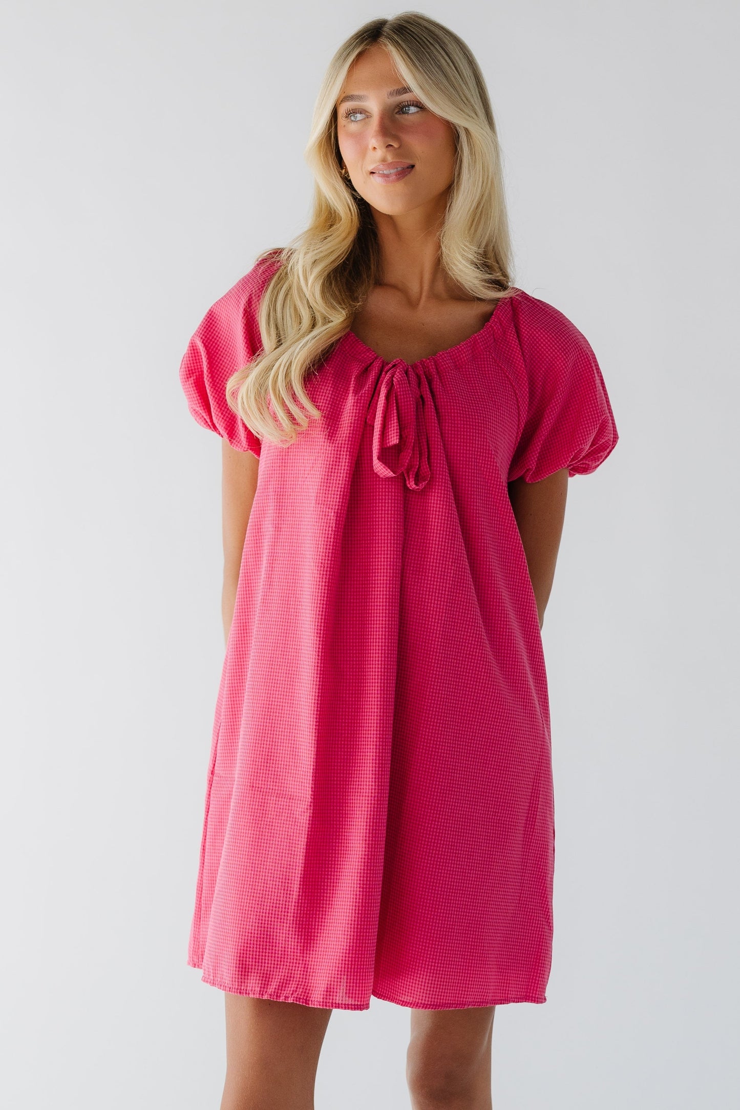 Young woman wearing a short red & pink gingham dress wtih short puff sleeves with a drawsting and bow scoop neckline.