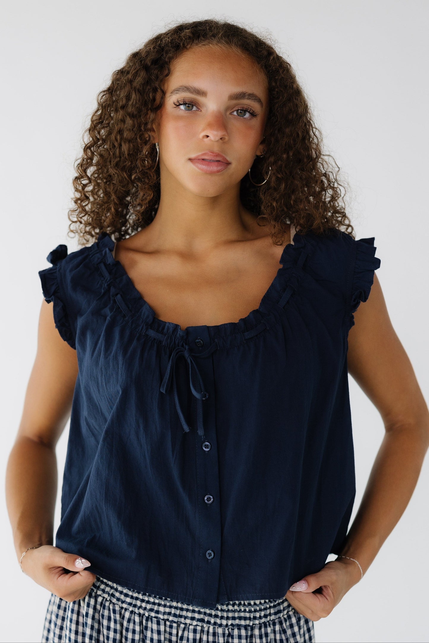 Teenage girl wearing a sleeveless button down blouse with ruffled arm openings, and a ruffled scoop neckline.