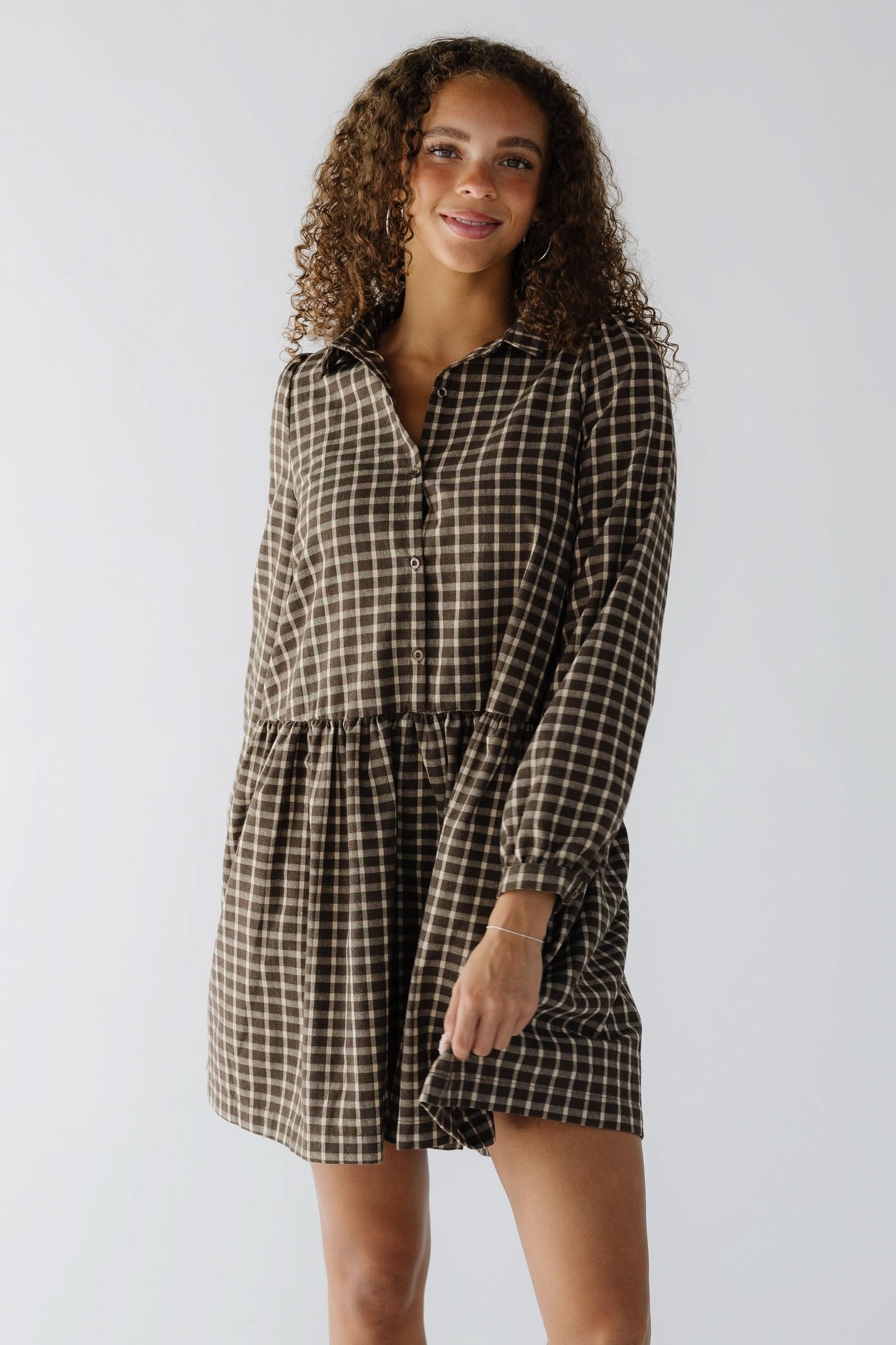 Woman wearing a  brown plaid short dress that buttons the front to the skirt, a pointed collar and long sleeves.