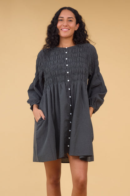 Young woman wearing a short dark grey gingham button down dress with a round neckline, spaced smocked bodice , long sleeves and side pockets.