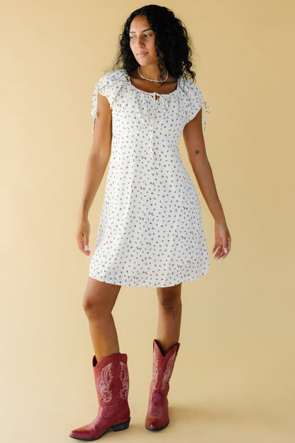 Young woman wearing a cream & red floral print knee length dress with a wide round neckline with a drawstring tie and puff sleeves.