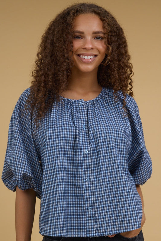 Teenage girl wearing a navy & blue gingham blouse with half length sleeves, round neckline and buttons down the front.