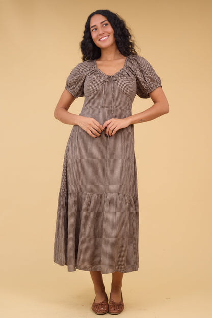 Young woman wearing a brown gingham midi dress with a wide bottom ruffle wide empire waist, short sleeves and drawstring neckline.