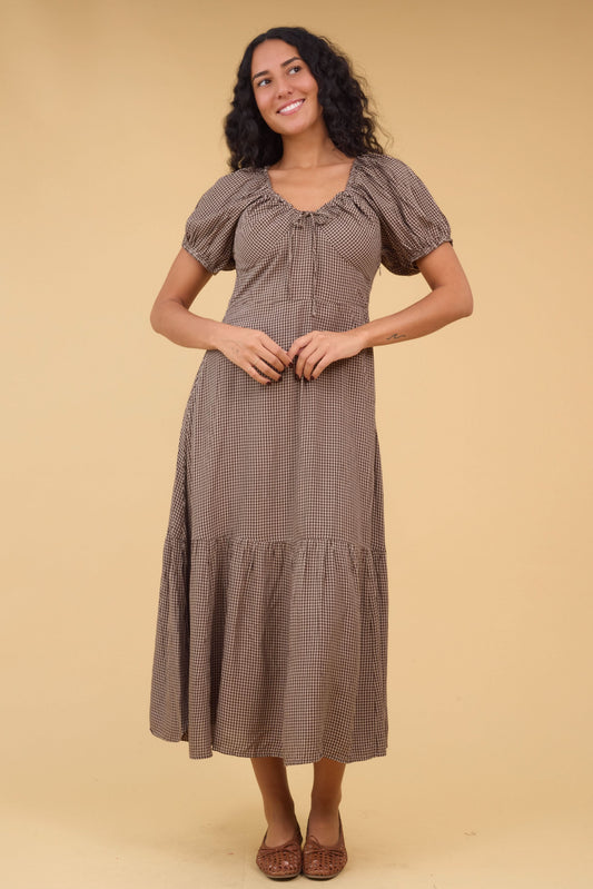 Young woman wearing a brown gingham midi dress with a wide bottom ruffle wide empire waist, short sleeves and drawstring neckline.