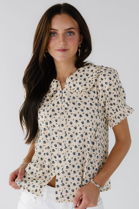 Young woman wearing a modest cream & blue print button down blouse with ruffle edged rounded collar and short puff sleeves.
