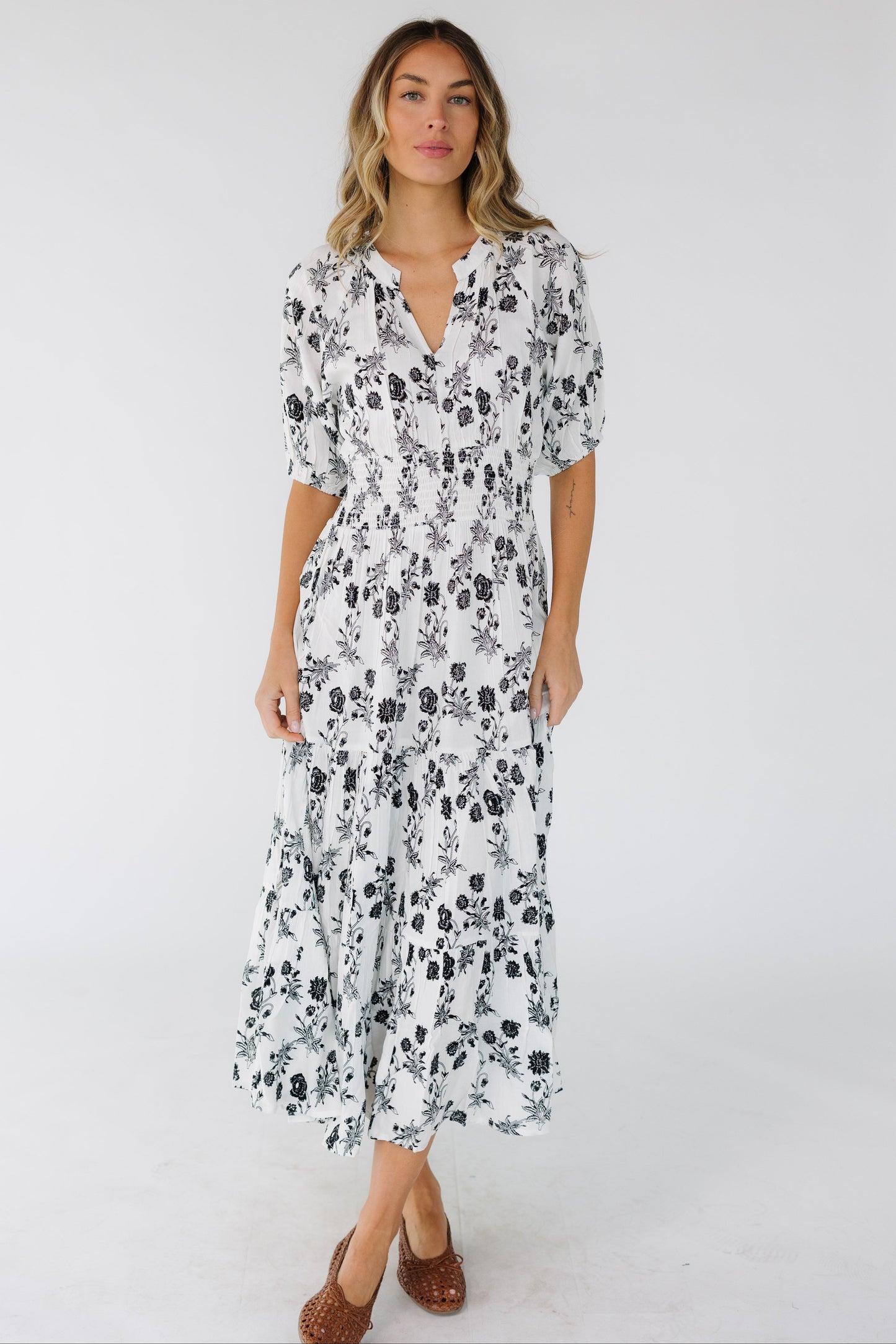 Woman wearing a white with black floral print midi dress with elbow length sleeves, a V-neckline and a wide elastic smocked waistband.