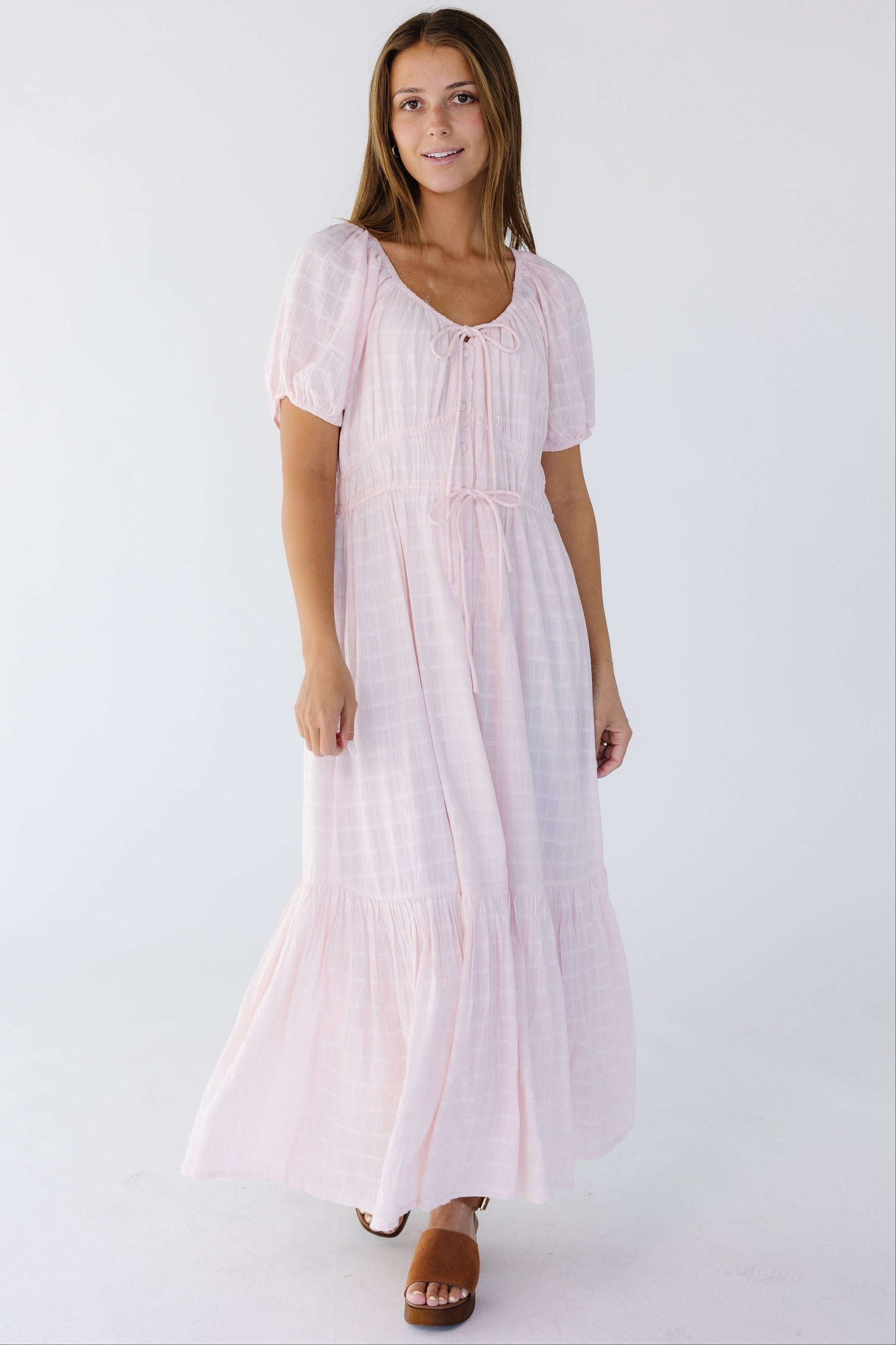 Young woman wearing a light pink midi-maxi dress with a textured plaid pattern with a scoop neckline, short puff sleeves and a full skirt.
