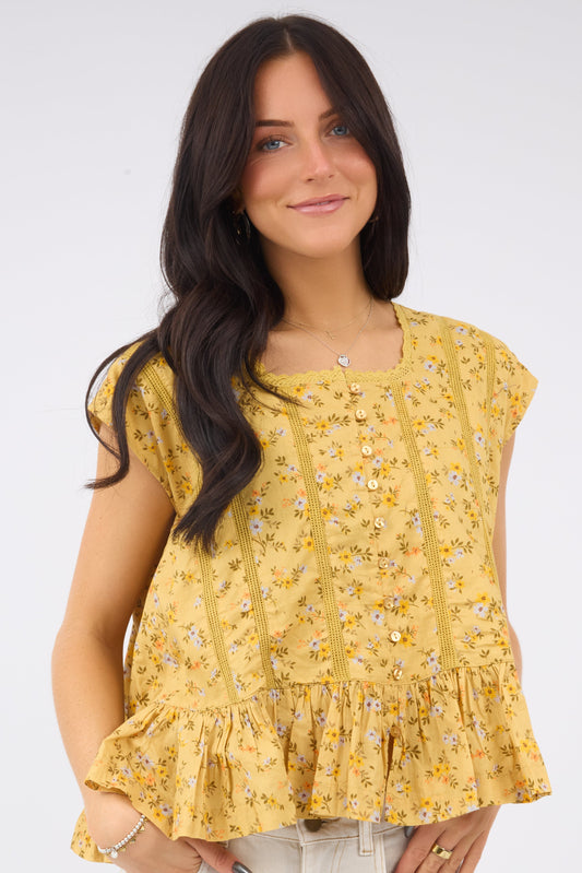 A young woman wearing a mustard yellow floral print blouse with cap sleeves, buttons with elstic loops and ruffle & lace details.
