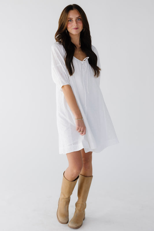 Young woman wearing a white eyelet short dress with a drawstring scoop neckline and elbow lenth paff sleeves.