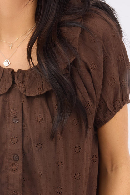 A close up view of a woman wearing a brown eyelet, button down blouse with short puff sleeve and a ruffled round neckline.