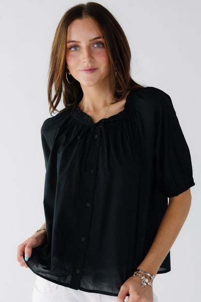 Young woman wearing a modest loose fitting button down top in black with short full sleeves and a stand up ruffle collar and tie.
