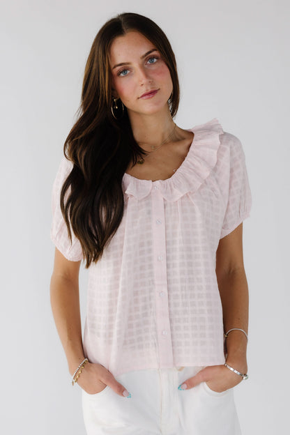Young woman wearing a light pink, plaid textured button down top with short sleeves and a ruffled collar.