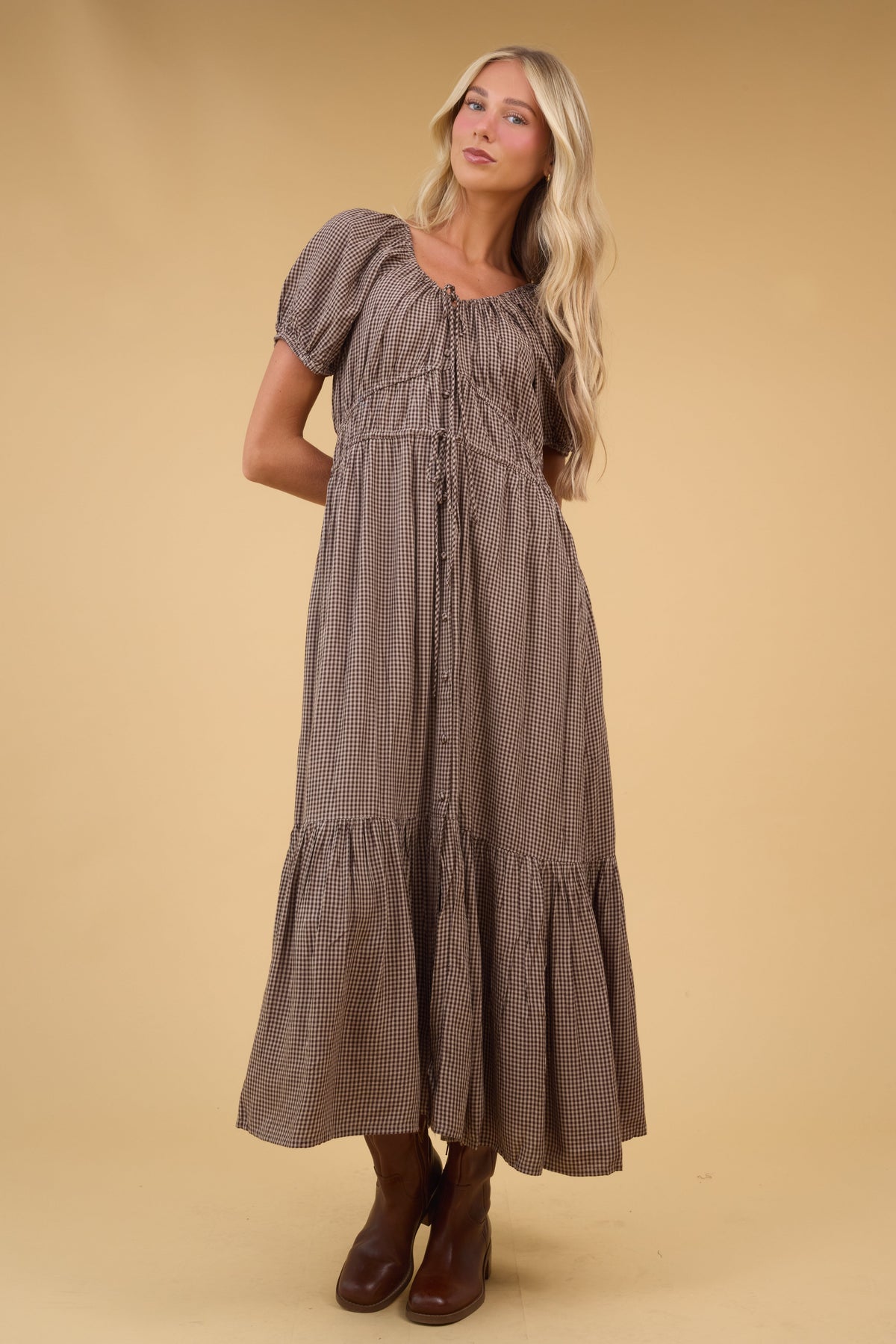 Young woman wearing a brown & white gingham midi dress with a drawstring scoop neckline, short puff sleeves and gathered-drawstring waist details with a wide bottom ruffle.