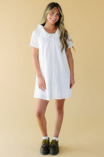 Young woman wearing a button down, white eyelet short dress with short puff sleeves and a round ruffled collar.