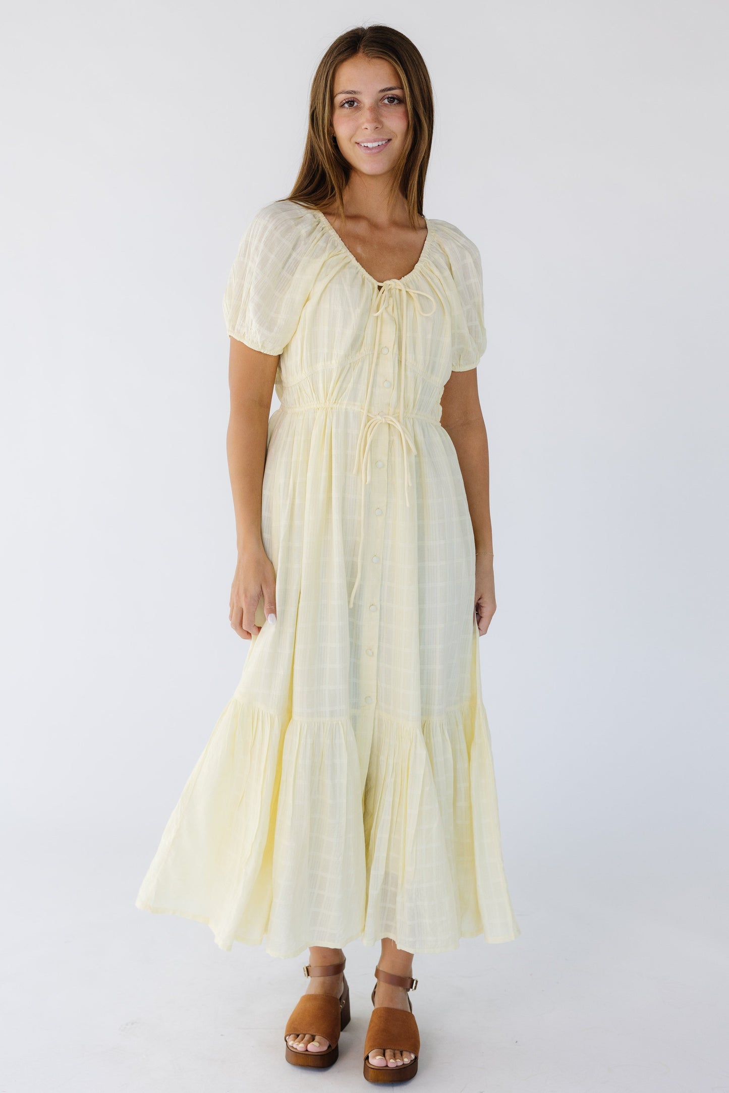Young woman wearing a light yellow button down midi-maxi dress with a drawstring scoop neckline and drawstring waist with a wide bottom ruffle
