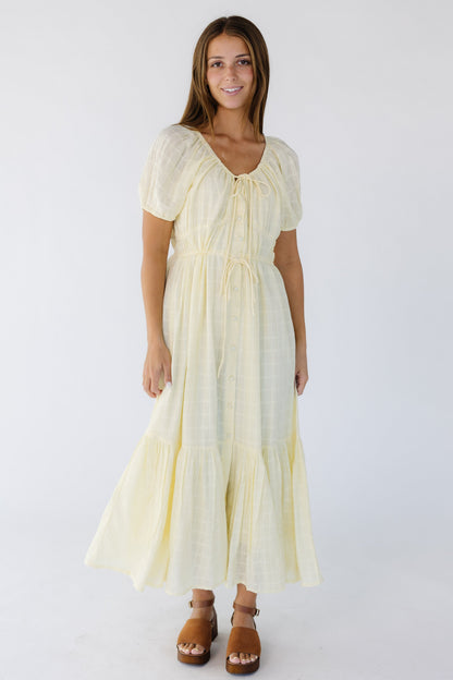 Young woman wearing a light yellow button down midi-maxi dress with a drawstring scoop neckline and drawstring waist with a wide bottom ruffle