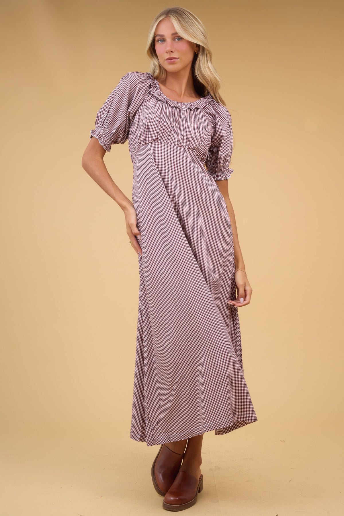 Young woman wearing a brown gingha midi dress with an empire waist, gathered bodice, short sleeves and a ruffled scoop neckline.