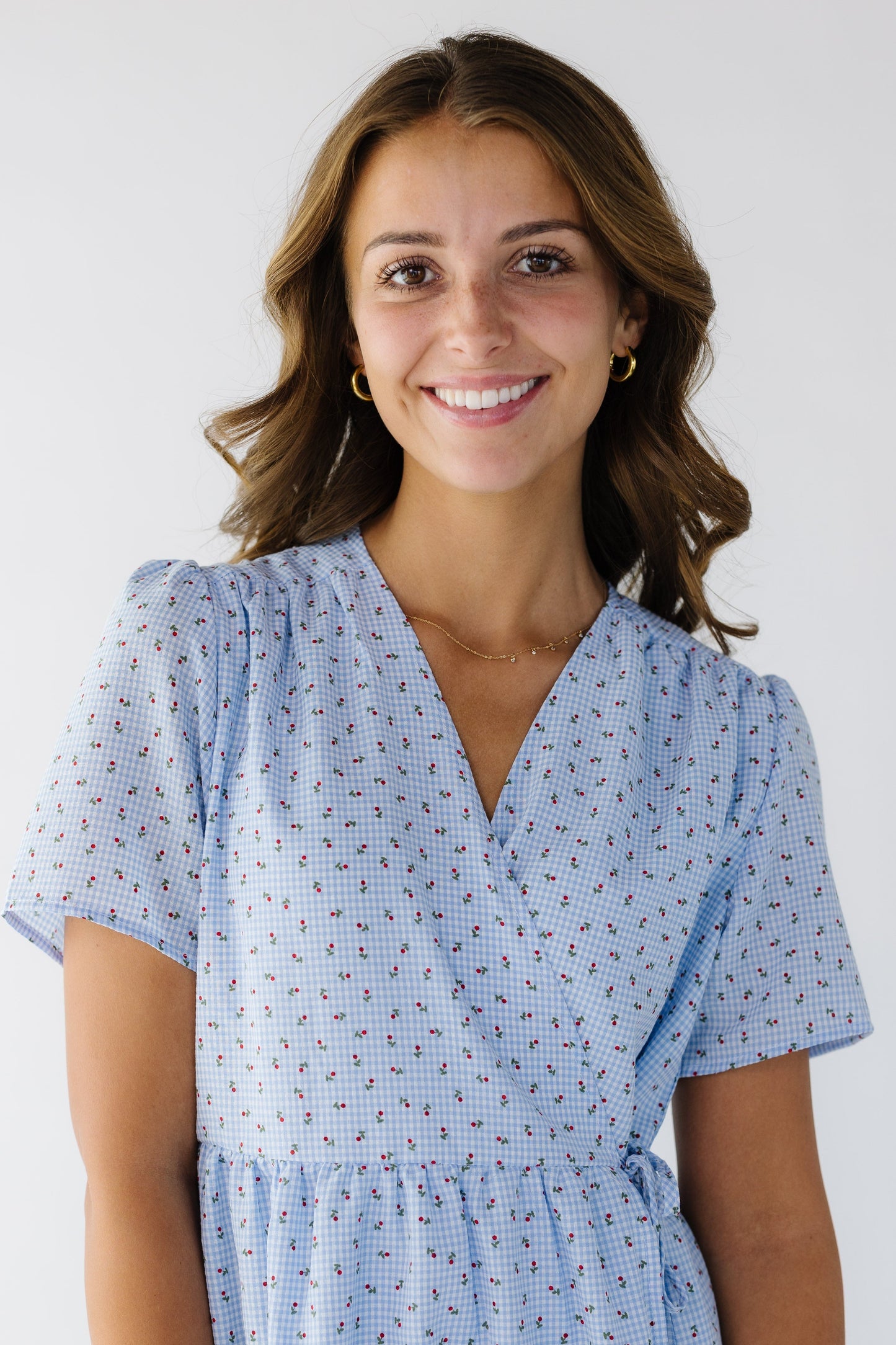 Close-up view of a young woman wearing a floral & light blue gingham wrap dress with short sleeves and a V-neckline.