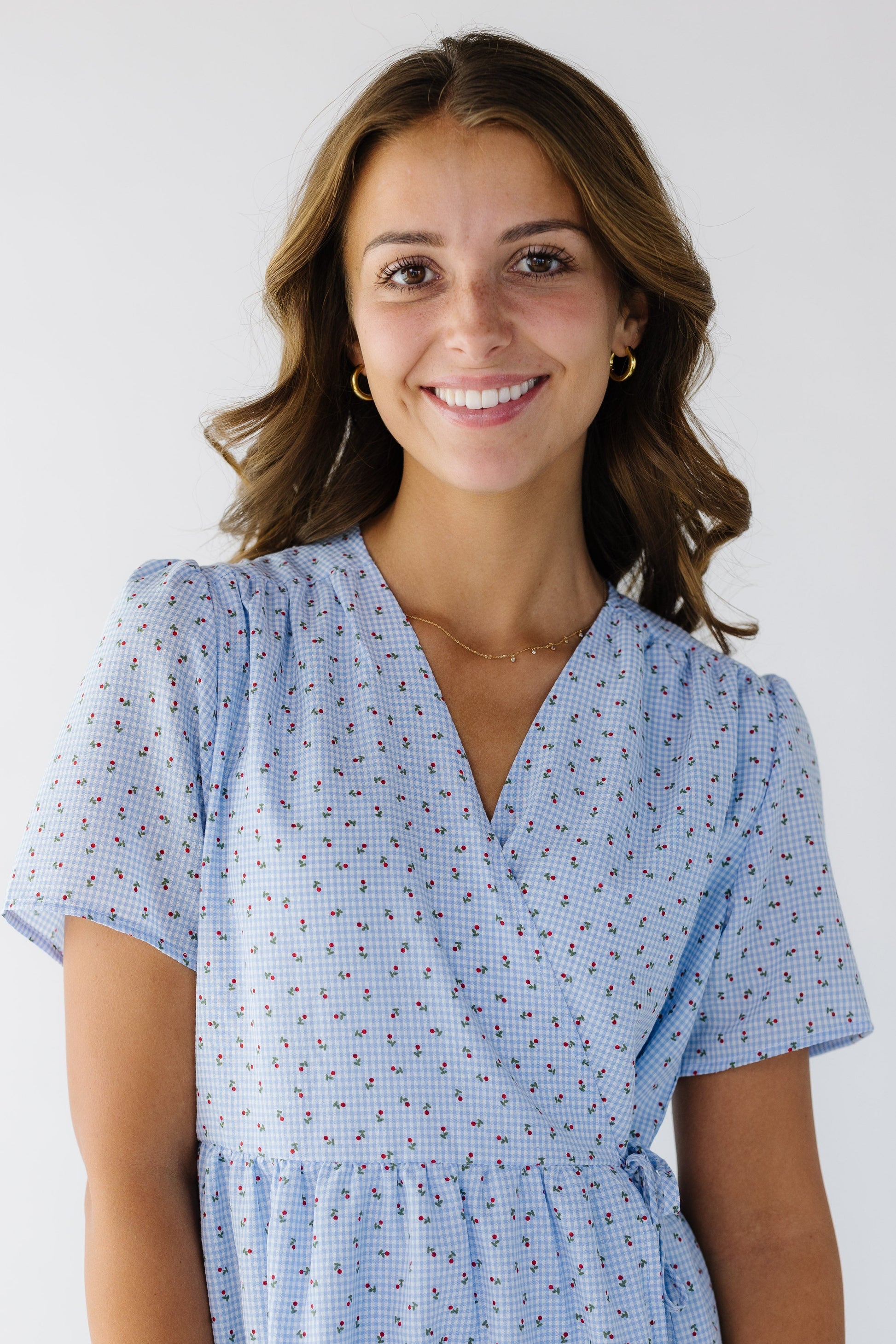 Close-up view of a young woman wearing a floral & light blue gingham wrap dress with short sleeves and a V-neckline.