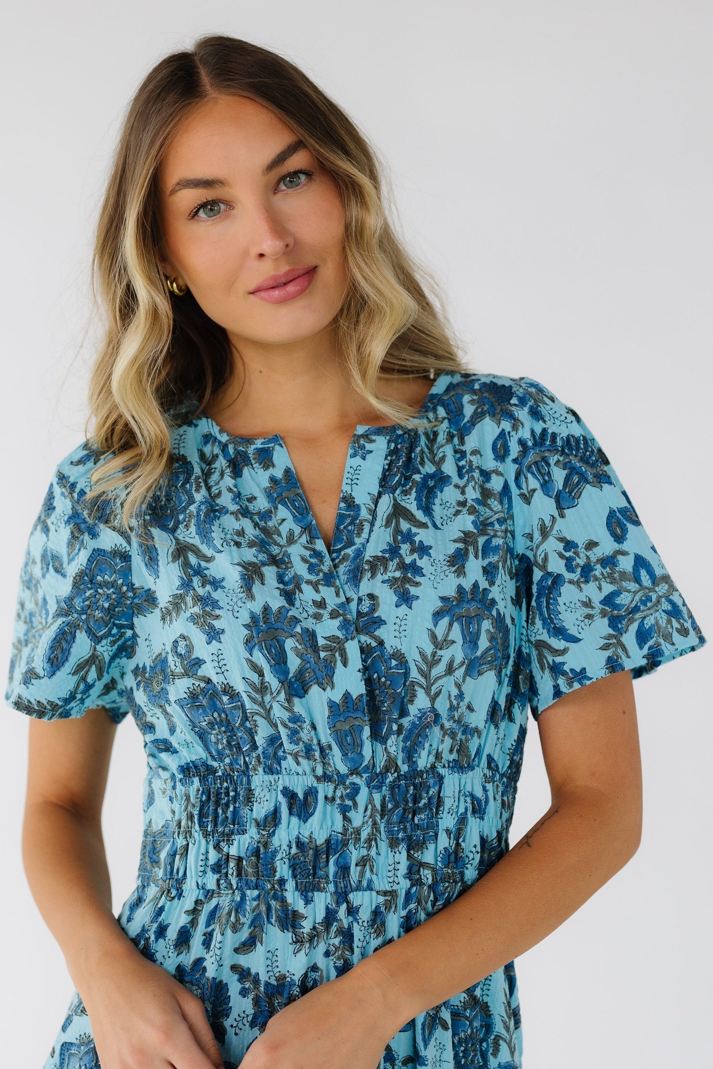 Young woman wearing a blue floral & paisley dress with a V-neckline, short sleeves and a wide elastic waist.