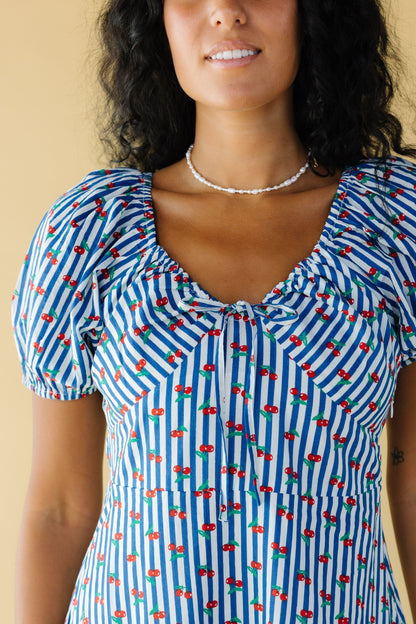 Close view of a woman wearing a cherry print & blue & white stripe dress with a close fitting empire waist, close fitting bodice and and large puff sleeves.