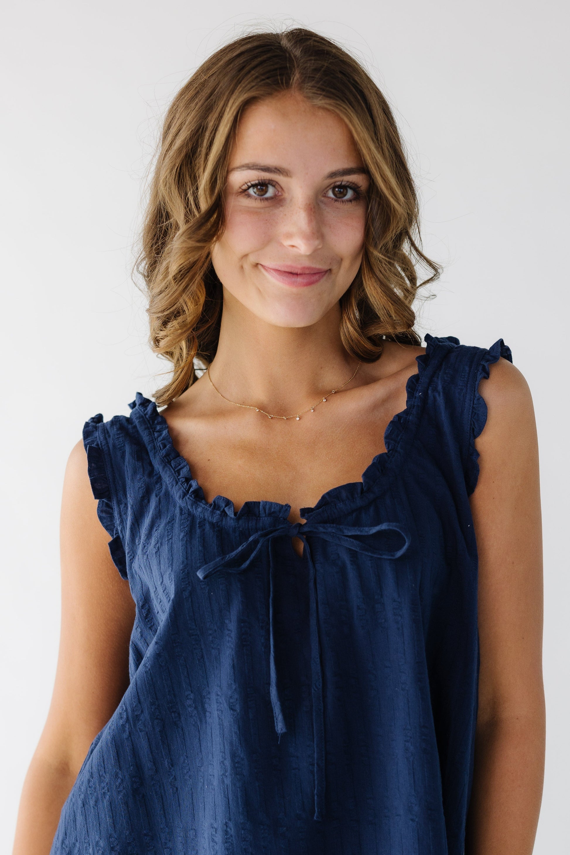 Close view of a young woman wearing a blue textured, sleeveless dress with a scoop neckline and ruffled neckline and around arm holes.