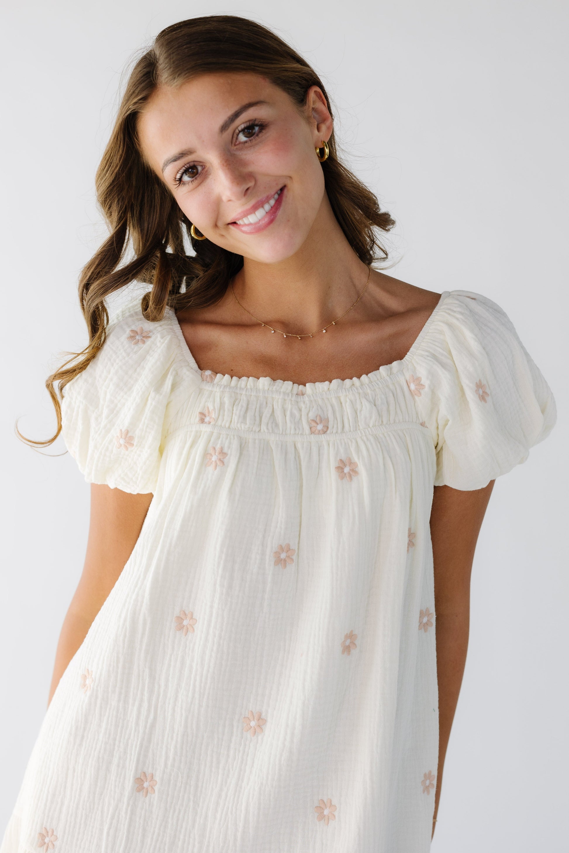 Close view of a young woman wearing a loose fitting swing dress in ivory with scattered pink embroidered daisy's, a square neckline and short puff sleeves.