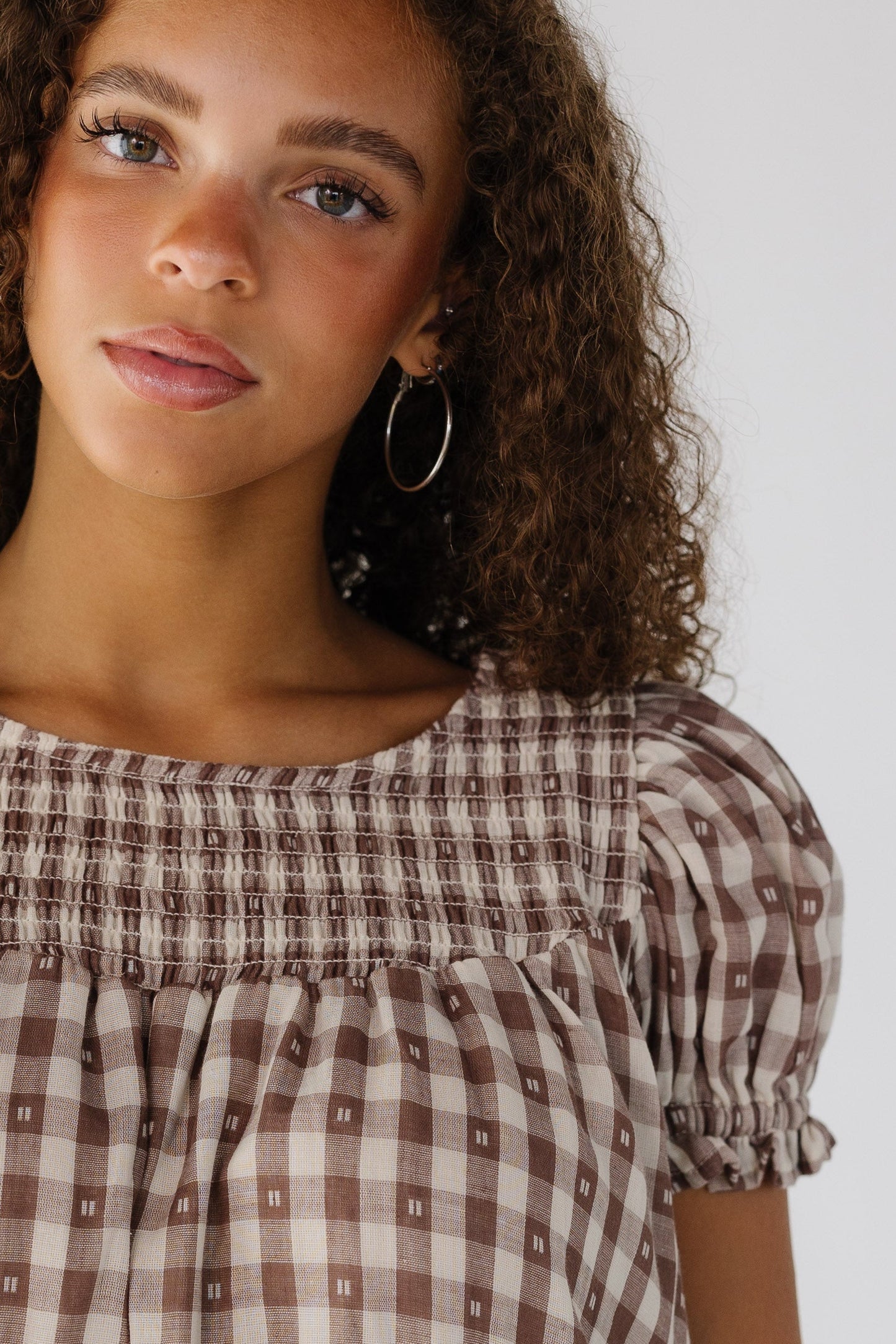 Close view of a young woman wearing a brown ginghamdress with puff sleeves and a smocked yoke.