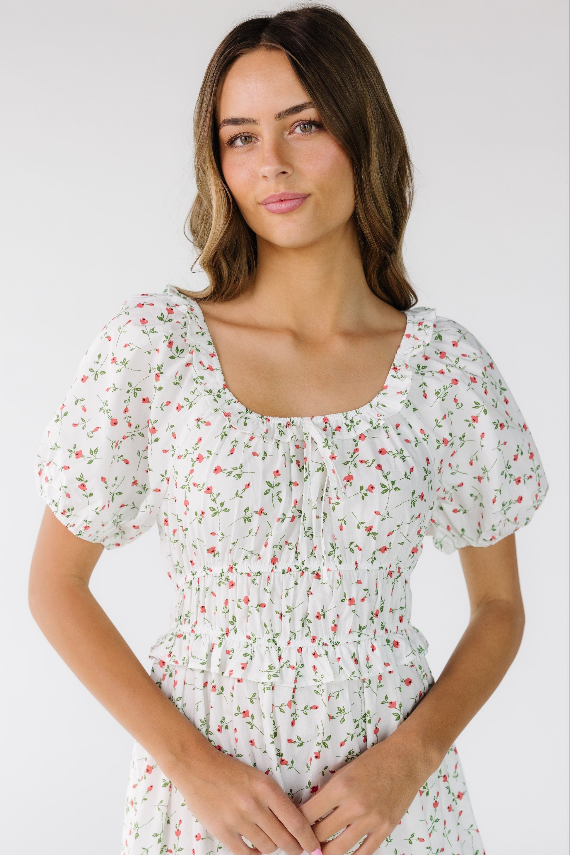 Close view of a young woman wearing an ivory and red floral print dress with a scoop neckline with a narrow ruffle, short puff sleeves and a wide close fitting waist.
