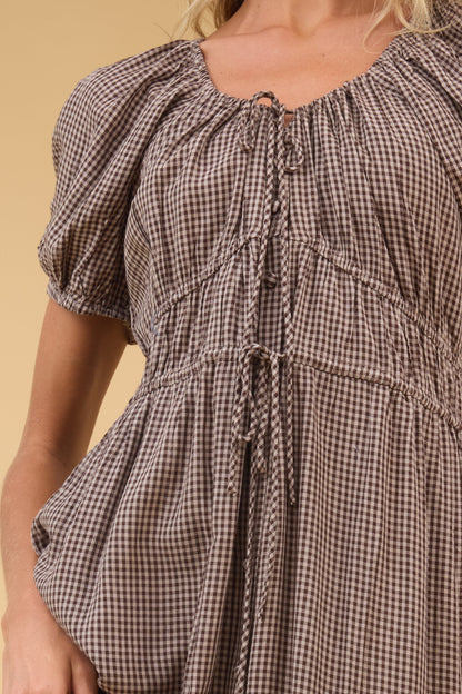 Close view of a woman wearing a brown gingham dress with drawstring-gathered waist details and short puff sleeves.
