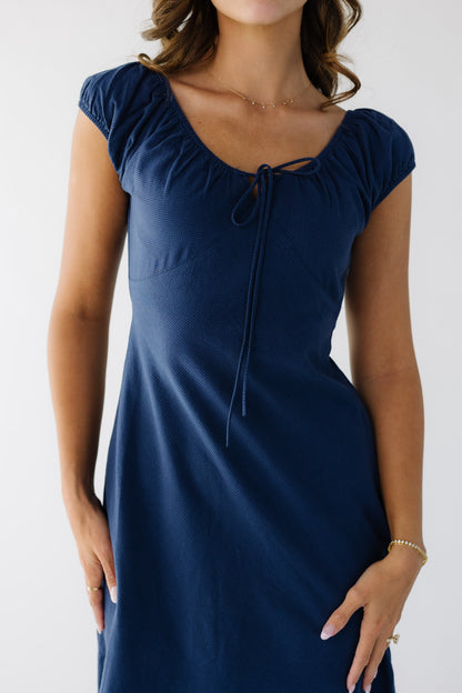 Close up view of a woman wearing a navy close fitting dress with a scoop neckline, empire waist and short puff sleeves.