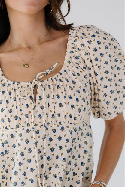 Close view of a woman wearing a cream and blue floral print dress with an empire waist, drawstring scoop neckline and balloon sleeves.
