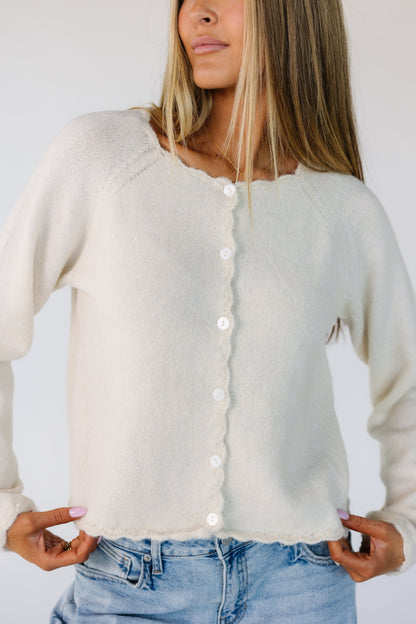 Close view of a woman wearing a cream button down cardigan with scallop edged neckline, cuffs, hems and button placket.