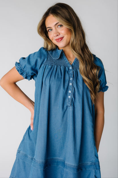 Woman wearing a light denim dress with a short button placket, smocked yoke and short puff sleeves.