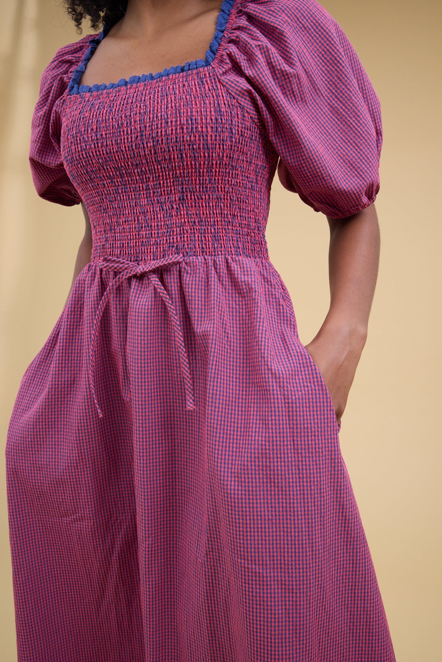 Close view of a woman wearing a blue & red gingham dress with balloon sleees, lacee edged square neckline, smocked bodice and side pockets.