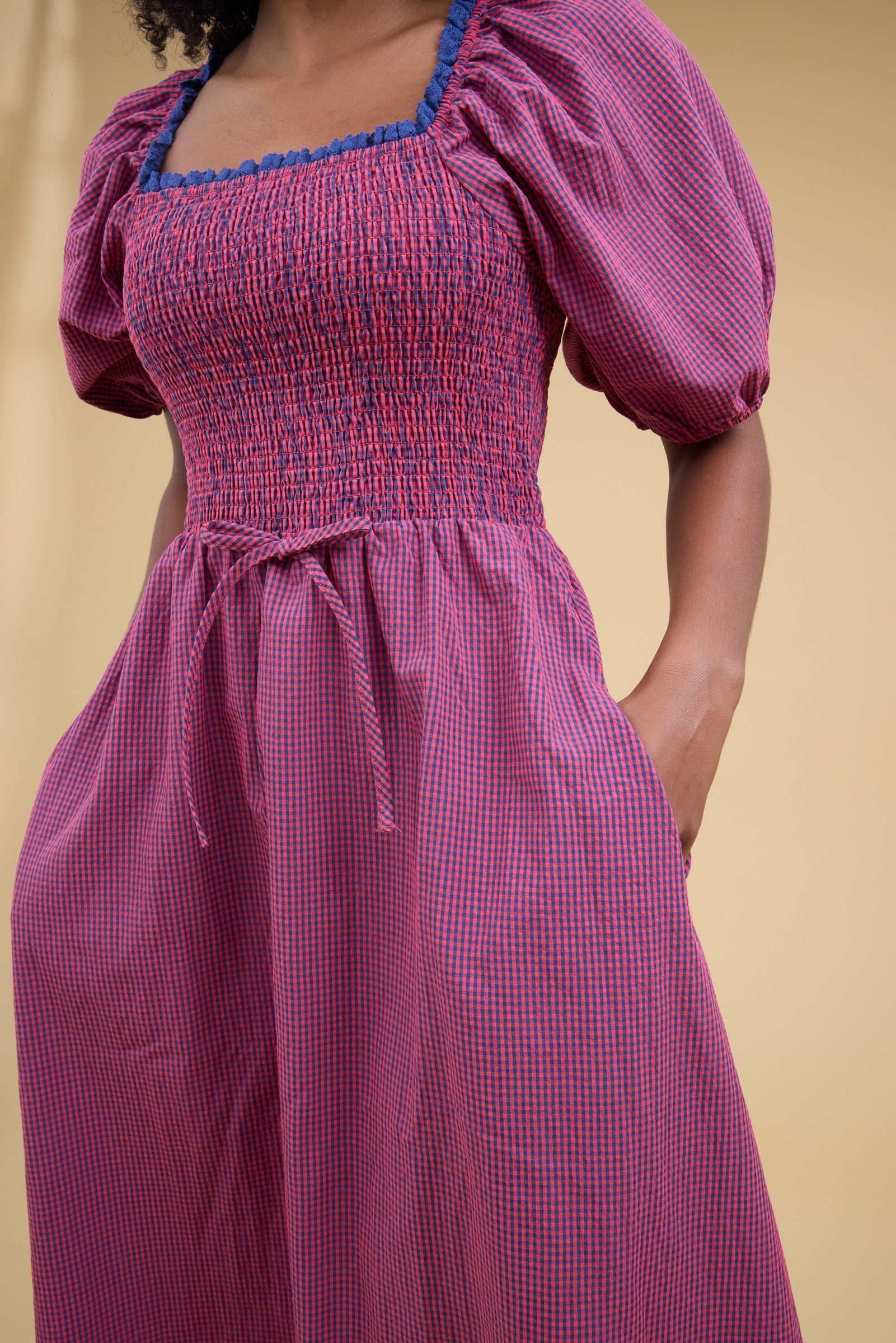 Close view of a woman wearing a blue & red gingham dress with balloon sleees, lacee edged square neckline, smocked bodice and side pockets.