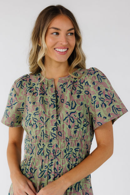 Close view of a woman wearing a green floral print dress with a V-neckline, short sleeves and a wide elastic waistband.