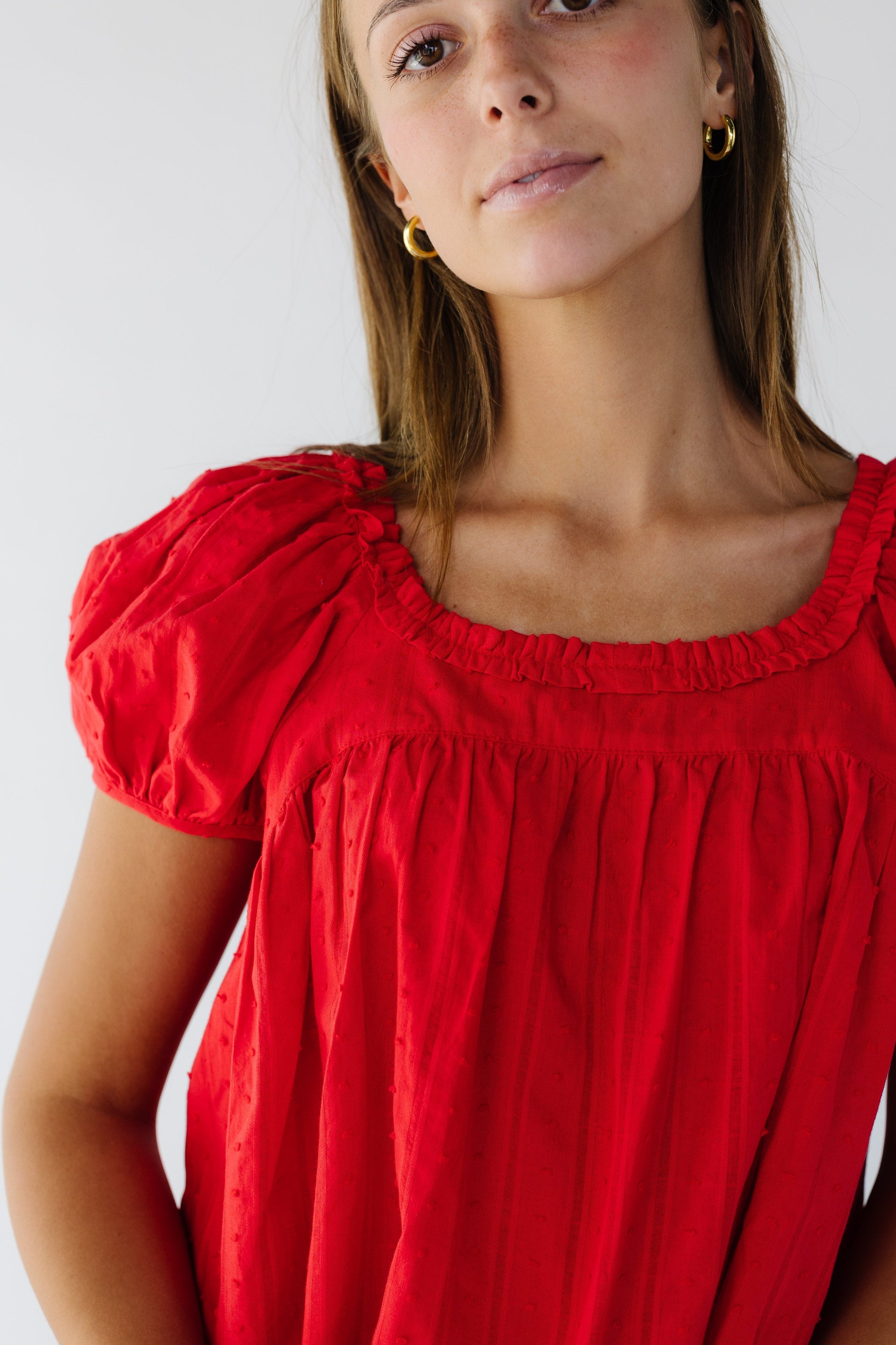 Close view of a young woman wearing a red loose fitting Swiss dot fabric with a ruffled square neckline and short puff sleeves.