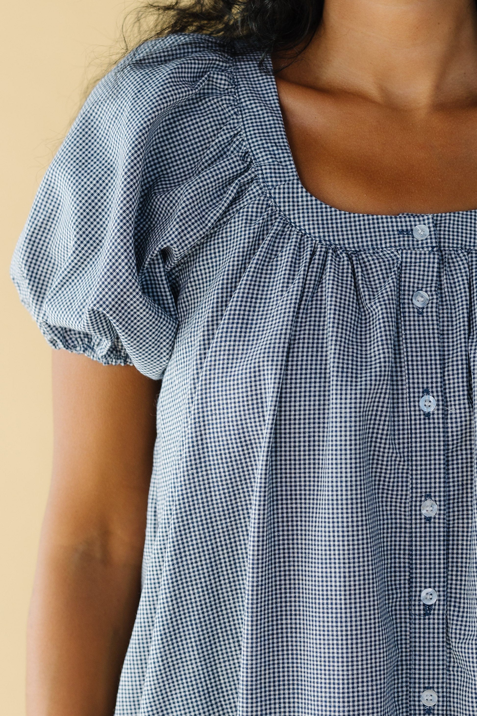 Close view of a woman wearing a navy & white gingham button down dress with a scoop neckline and large puff sleeves.